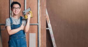 A person wearing glasses and denim overalls stands indoors, holding a nail gun, with construction materials and unfinished walls in the background—capturing the expertise often seen with a Boon Woh fire rated door contractor Singapore during the installation process.