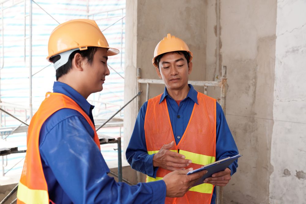 Two construction workers in blue uniforms and orange safety vests discuss a project while looking at a tablet inside a building under construction, reviewing details provided by the Boon Woh fire rated door contractor Singapore for enhanced site safety and compliance.