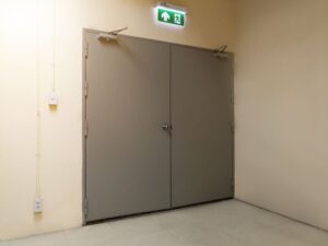 Closed double doors in a beige hallway with an illuminated green emergency exit sign above, featuring a Boon Woh fire rated door in Singapore, and visible wall outlets and switches nearby.