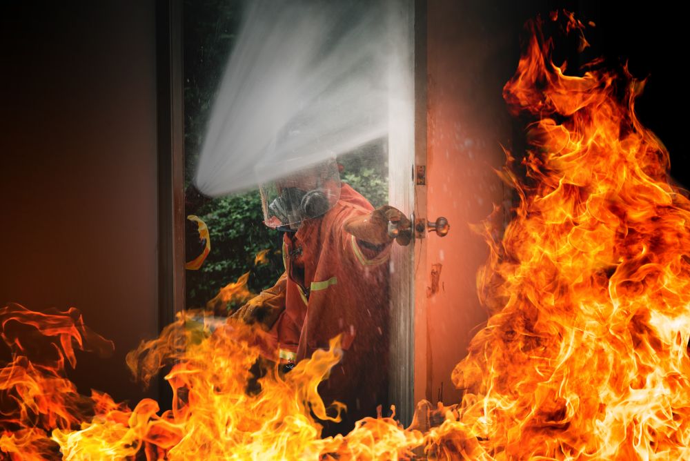 A firefighter sprays water through a Boon Woh fire rated door in Singapore, directing the stream into a burning room surrounded by intense flames and thick smoke.