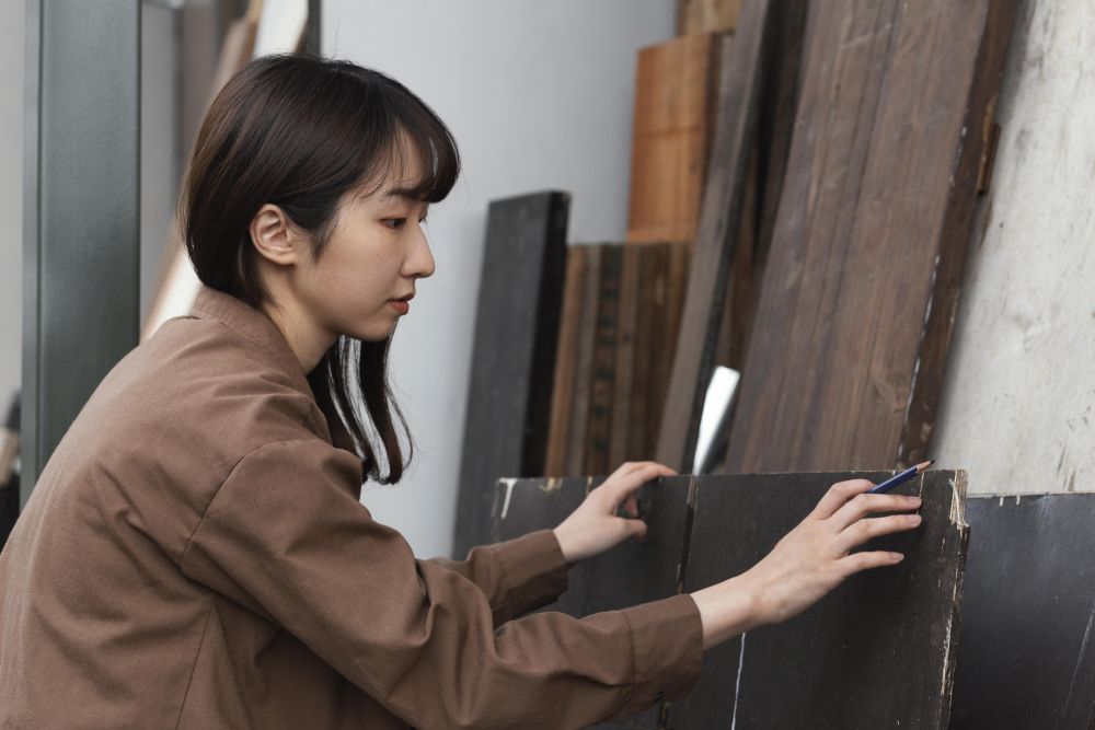 A person wearing a brown shirt arranges dark wooden boards while holding a pencil, positioned in front of a vertical stack of wood panels, possibly preparing a Boon Woh timber fire rated door.