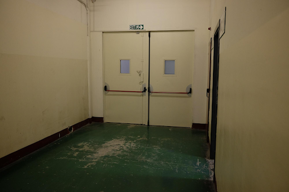Empty hallway with green floor, beige walls, and double doors at the end; a commercial fire rated door by Boon Woh Singapore is installed beneath the exit sign above the doors.